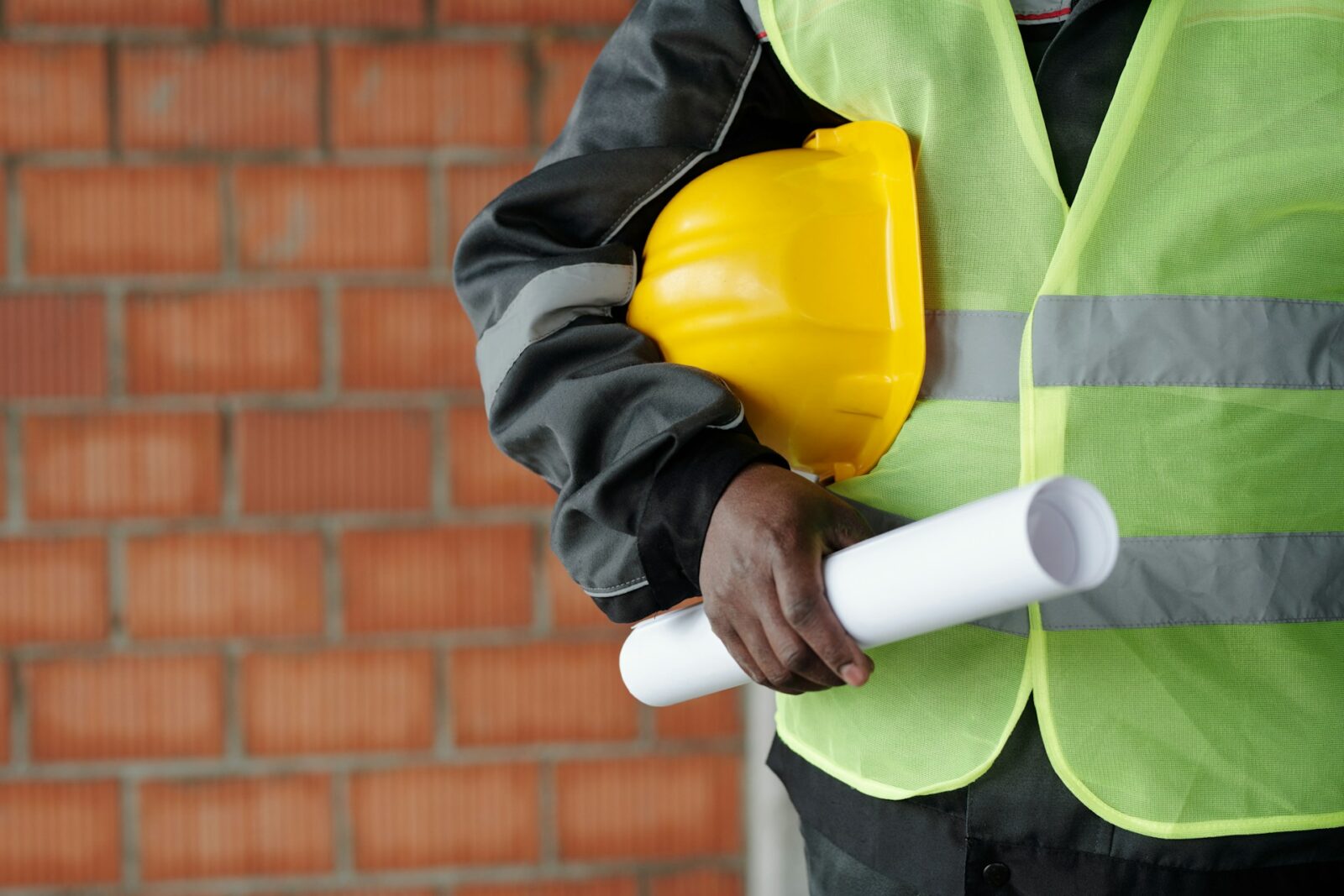 onstruction worker holding a yellow helmet and a rolled-up blueprint, wearing a high-visibility safety vest in front of a brick wall.