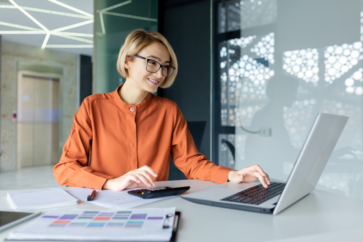 Office worker smiling while multitasking with a laptop, calculator, and documents.