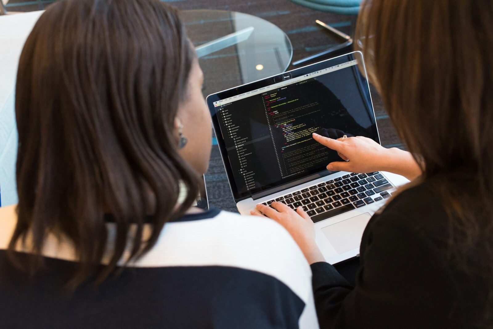 Two women collaborating on coding, with one pointing at a laptop screen displaying lines of code